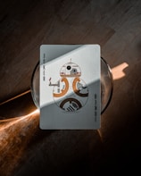 A playing card featuring a character resembling a robot is placed on a wooden surface. The robot design on the card appears in shades of orange, brown, and white. Light and shadow create a diagonal line across the card, adding contrast and highlighting its surface texture.