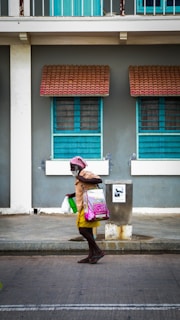 An elderly person walks along a street carrying several bags, one of which is pink and another white. The individual wears a yellow skirt, brown top, and a headscarf. The background features a building with two turquoise-colored windows and red-tiled awnings.