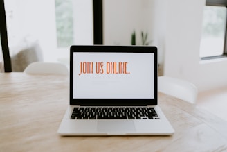A laptop is placed on a wooden table in a bright room. The screen displays the text 'JOIN US ONLINE.' in bold orange letters. The surroundings are minimalistic with soft lighting, suggesting a comfortable and modern environment.