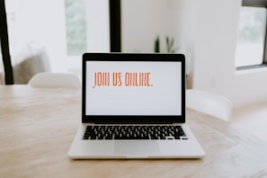 A laptop is placed on a wooden table in a bright room. The screen displays the text 'JOIN US ONLINE.' in bold orange letters. The surroundings are minimalistic with soft lighting, suggesting a comfortable and modern environment.