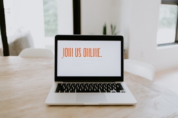 A laptop is placed on a wooden table in a bright room. The screen displays the text 'JOIN US ONLINE.' in bold orange letters. The surroundings are minimalistic with soft lighting, suggesting a comfortable and modern environment.