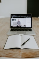 A laptop displaying a video of a person sitting on a couch is seen on a wooden table. In front of the laptop, there is an open notebook with a pen placed on it.