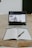 A laptop displaying a video of a person sitting on a couch is seen on a wooden table. In front of the laptop, there is an open notebook with a pen placed on it.