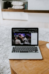 A laptop displaying a screen with a group of people is placed on a round wooden table. In the background, there are shelves with neatly arranged books and other items. The setting appears to be a comfortable, relaxed indoor environment with a soft carpet.