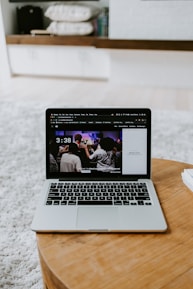 A laptop displaying a screen with a group of people is placed on a round wooden table. In the background, there are shelves with neatly arranged books and other items. The setting appears to be a comfortable, relaxed indoor environment with a soft carpet.
