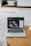 A laptop displaying a screen with a group of people is placed on a round wooden table. In the background, there are shelves with neatly arranged books and other items. The setting appears to be a comfortable, relaxed indoor environment with a soft carpet.