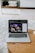 A laptop displaying a screen with a group of people is placed on a round wooden table. In the background, there are shelves with neatly arranged books and other items. The setting appears to be a comfortable, relaxed indoor environment with a soft carpet.