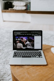 A laptop displaying a screen with a group of people is placed on a round wooden table. In the background, there are shelves with neatly arranged books and other items. The setting appears to be a comfortable, relaxed indoor environment with a soft carpet.