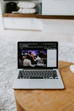 A laptop displaying a screen with a group of people is placed on a round wooden table. In the background, there are shelves with neatly arranged books and other items. The setting appears to be a comfortable, relaxed indoor environment with a soft carpet.