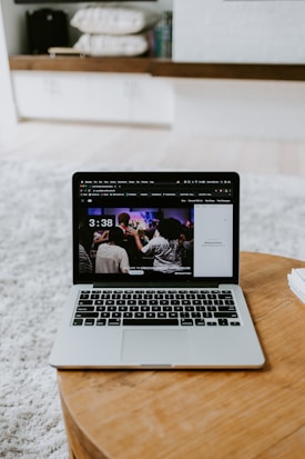 A laptop displaying a screen with a group of people is placed on a round wooden table. In the background, there are shelves with neatly arranged books and other items. The setting appears to be a comfortable, relaxed indoor environment with a soft carpet.