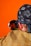 A person wearing a checkered jacket and a bandana with intricate patterns is looking into a small round mirror. The bandana features symbols and designs, possibly of cultural significance. The background is bright orange, enhancing the image's vibrancy.