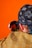 A person wearing a checkered jacket and a bandana with intricate patterns is looking into a small round mirror. The bandana features symbols and designs, possibly of cultural significance. The background is bright orange, enhancing the image's vibrancy.
