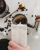 A cup of coffee is placed on a white surface, surrounded by scattered coffee beans. A hand is holding a carton, and there's a gold-colored scoop nearby. A glass carafe with a wooden collar is partially visible, suggesting a coffee preparation scene.