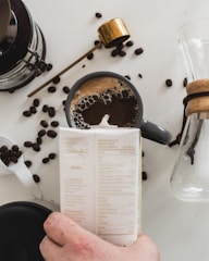 A cup of coffee is placed on a white surface, surrounded by scattered coffee beans. A hand is holding a carton, and there's a gold-colored scoop nearby. A glass carafe with a wooden collar is partially visible, suggesting a coffee preparation scene.