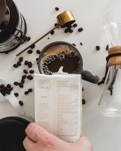 A cup of coffee is placed on a white surface, surrounded by scattered coffee beans. A hand is holding a carton, and there's a gold-colored scoop nearby. A glass carafe with a wooden collar is partially visible, suggesting a coffee preparation scene.