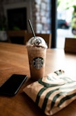 brown and white starbucks cup beside black smartphone on brown wooden table