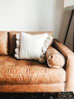 A tan leather sofa with a plush, white pillow in the center. The pillow has decorative, fluffy pom-poms around its edges. On the right side, an armrest rounds off smoothly, and in the background, a tall floor lamp with a tripod base is partially visible against a plain white wall.