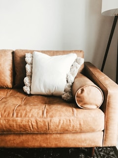 A tan leather sofa with a plush, white pillow in the center. The pillow has decorative, fluffy pom-poms around its edges. On the right side, an armrest rounds off smoothly, and in the background, a tall floor lamp with a tripod base is partially visible against a plain white wall.