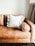 A tan leather sofa with a plush, white pillow in the center. The pillow has decorative, fluffy pom-poms around its edges. On the right side, an armrest rounds off smoothly, and in the background, a tall floor lamp with a tripod base is partially visible against a plain white wall.