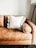 A tan leather sofa with a plush, white pillow in the center. The pillow has decorative, fluffy pom-poms around its edges. On the right side, an armrest rounds off smoothly, and in the background, a tall floor lamp with a tripod base is partially visible against a plain white wall.