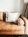 A tan leather sofa with a plush, white pillow in the center. The pillow has decorative, fluffy pom-poms around its edges. On the right side, an armrest rounds off smoothly, and in the background, a tall floor lamp with a tripod base is partially visible against a plain white wall.