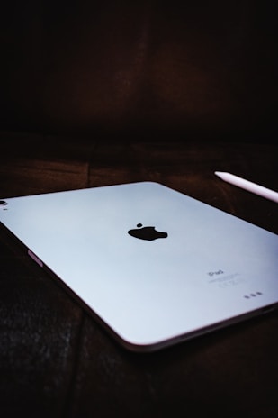 A close-up view of a sleek tablet with a metallic finish, prominently displaying a black apple logo in the center. The device is slightly angled on a dark surface, accompanied by a white stylus placed nearby.