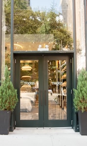 A retail store entrance with glass double doors, reflecting the surroundings. Flanking the doors are two tall potted plants. Inside, various home textiles such as pillows and sheets are visible. The store has a modern interior with neutral tones.
