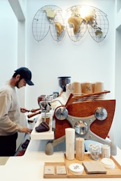 A barista is preparing coffee using a modern espresso machine with a wooden panel. The counter is organized with coffee cups, stirrers, napkins, and coasters, each printed with a percentage symbol. Above the barista, there's a decorative piece featuring a world map made of metal.