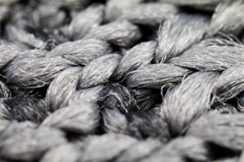 A close-up view of intricately woven gray fibers creating a thick, textured pattern. The strands exhibit a glossy sheen, highlighting the twisting arrangement.