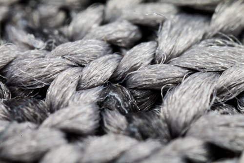 A close-up view of intricately woven gray fibers creating a thick, textured pattern. The strands exhibit a glossy sheen, highlighting the twisting arrangement.