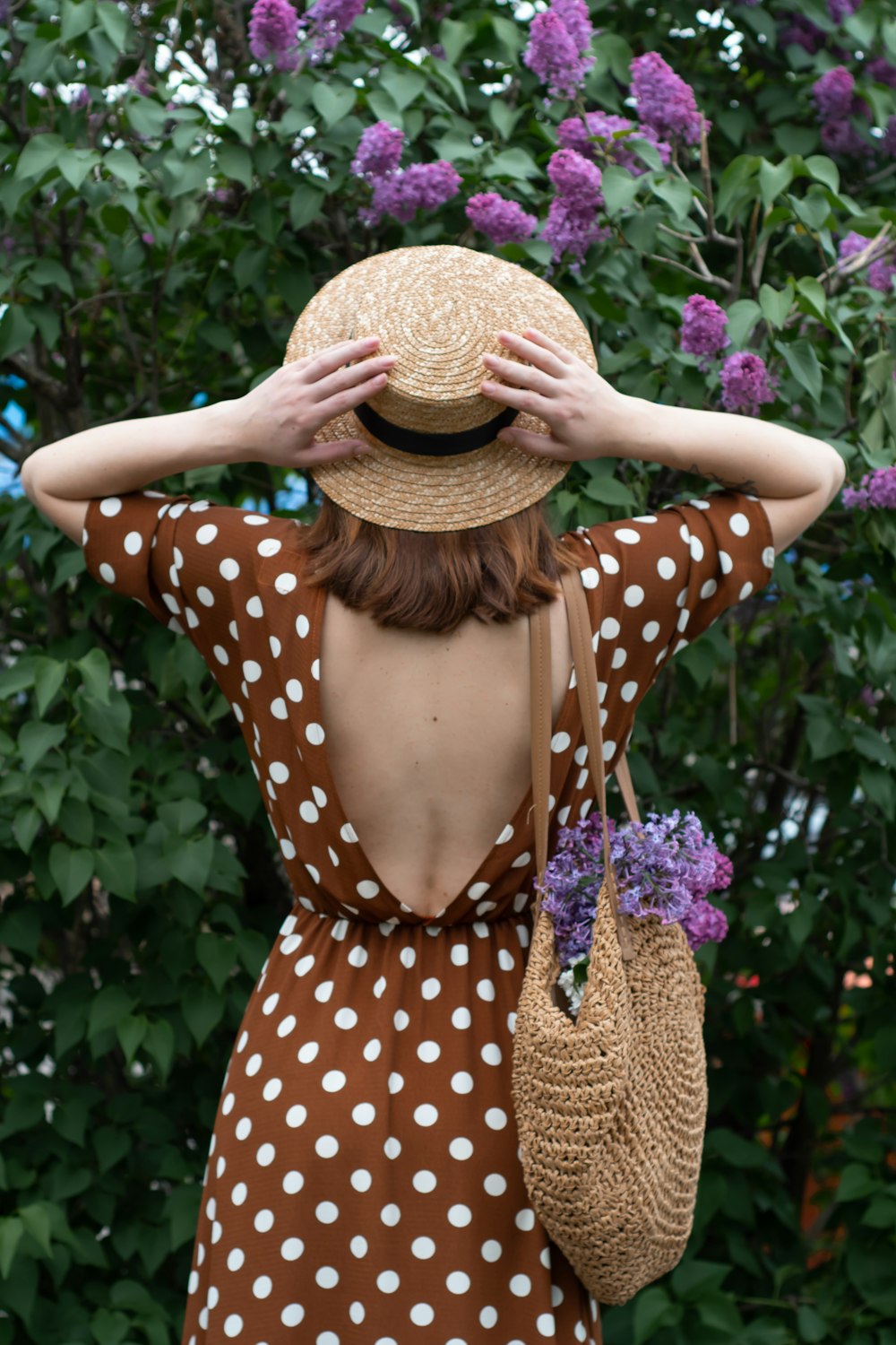woman in white and black polka dot dress wearing brown straw hat standing near purple flowers