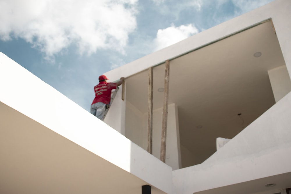 Mann in roter Jacke steht auf weißem Betongebäude unter blau-weißem Wolkenhimmel während