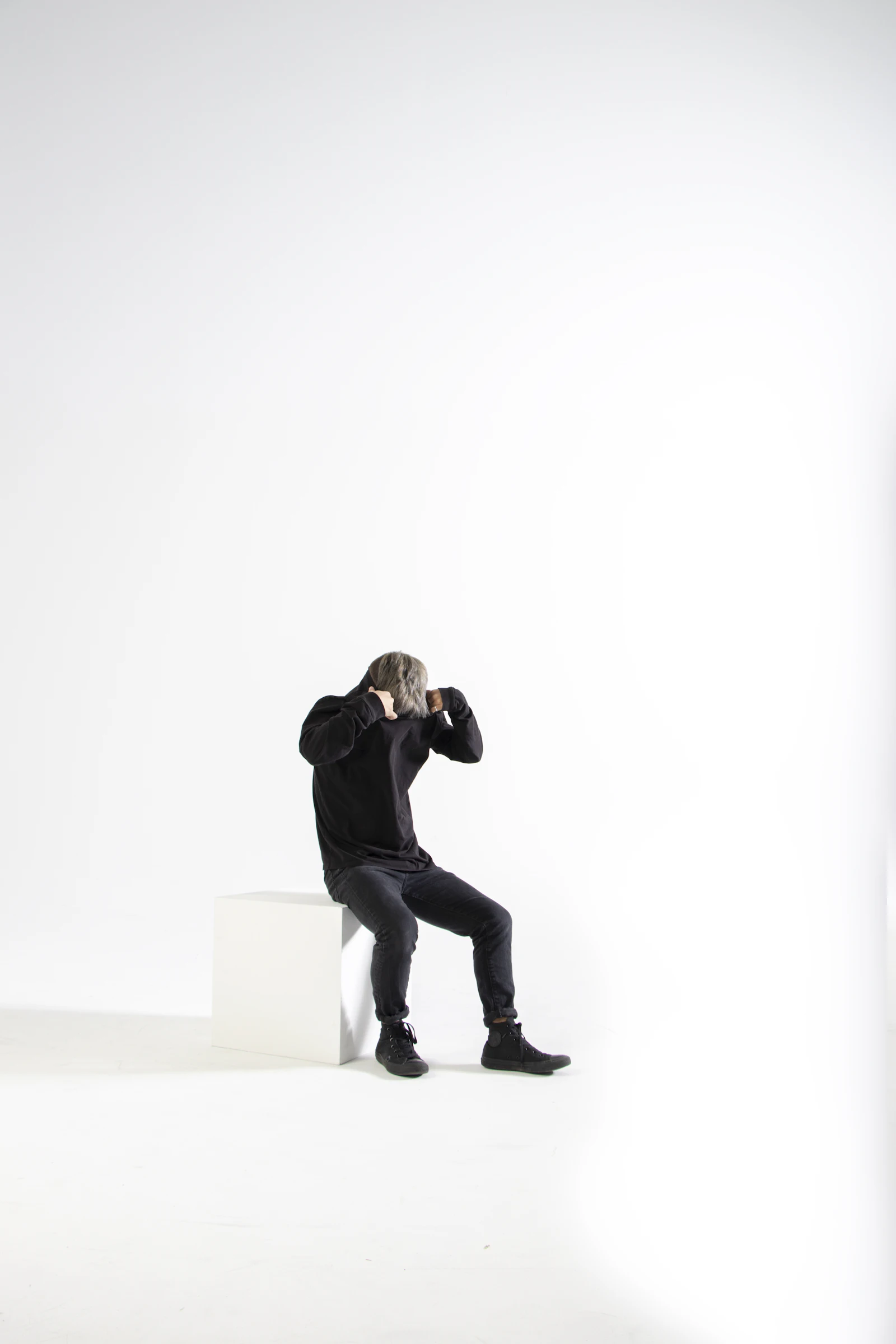 Model in a black hoodie and black pants sitting on a white block in a white studio