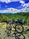 A mountain bike is parked on a rocky trail surrounded by lush green trees. In the background, a vast expanse of forest stretches out under a bright blue sky dotted with fluffy white clouds. The scene suggests a peaceful and adventurous outdoor setting.