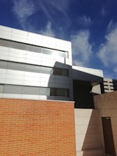Modern architectural building featuring a mix of materials, including a high brick wall and a sleek, metallic facade. The structure has a geometric design with clean lines and large reflective windows. The sky above is clear and deep blue, with a few wispy clouds casting shadows on the building.