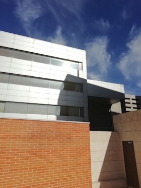 Modern architectural building featuring a mix of materials, including a high brick wall and a sleek, metallic facade. The structure has a geometric design with clean lines and large reflective windows. The sky above is clear and deep blue, with a few wispy clouds casting shadows on the building.