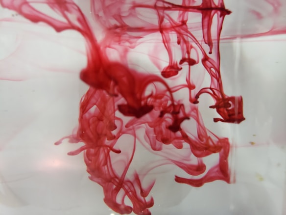 Red ink flows and swirls in clear water, creating abstract shapes and patterns. The ink appears to spread in various directions with wispy tendrils and cloud-like formations.