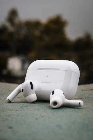 A pair of wireless earbuds and their charging case are positioned on a surface outdoors. The earbuds are white with black accents, and the charging case is also white, displaying some text. The background appears blurred, with trees in muted colors.