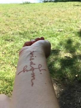 A person's arm is extended with a temporary reddish-brown tattoo of the word 'wisdom' written vertically. The background features a grassy area with patches of sunlight and shade.