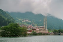 A picturesque landscape featuring a large mosque with two tall minarets near a tranquil lake, surrounded by lush greenery and distant hills partially obscured by clouds. A small town is nestled amongst the foothills, offering a serene and peaceful setting.