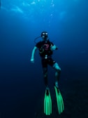 A scuba diver is underwater, equipped with diving gear including a mask, regulator, and fluorescent green fins. The surrounding water is deep and blue, with some light filtering down from above, creating a serene and calm atmosphere.