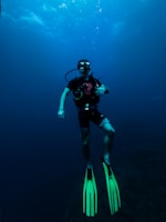 A scuba diver is underwater, equipped with diving gear including a mask, regulator, and fluorescent green fins. The surrounding water is deep and blue, with some light filtering down from above, creating a serene and calm atmosphere.