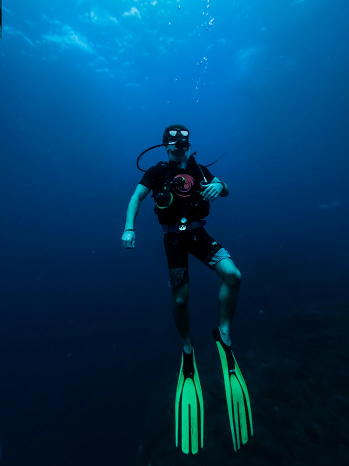 A scuba diver is underwater, equipped with diving gear including a mask, regulator, and fluorescent green fins. The surrounding water is deep and blue, with some light filtering down from above, creating a serene and calm atmosphere.