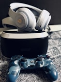 A pair of over-ear headphones rests on top of a virtual reality headset. Below them, a camo-patterned gaming controller is placed on a detailed, monochrome patterned surface. The arrangement appears organized and the lighting is soft, accentuating the sleek design of the gadgets.