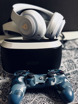 A pair of over-ear headphones rests on top of a virtual reality headset. Below them, a camo-patterned gaming controller is placed on a detailed, monochrome patterned surface. The arrangement appears organized and the lighting is soft, accentuating the sleek design of the gadgets.