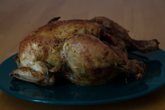 A roasted chicken with a golden brown, crispy skin is placed on a dark green plate. The background features a wooden table surface, creating a warm, home-cooked meal atmosphere.