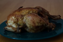 A roasted chicken with a golden brown, crispy skin is placed on a dark green plate. The background features a wooden table surface, creating a warm, home-cooked meal atmosphere.