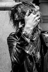 A person with an intense gaze is partially obscured by their hand, adorned with rings and bracelets. The individual wears a leather jacket and has long, textured hair, giving off a rock or punk aesthetic. Chains and jewelry add to the edgy look, and the photograph's black-and-white filter accentuates the mood.