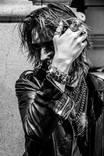 A person with an intense gaze is partially obscured by their hand, adorned with rings and bracelets. The individual wears a leather jacket and has long, textured hair, giving off a rock or punk aesthetic. Chains and jewelry add to the edgy look, and the photograph's black-and-white filter accentuates the mood.