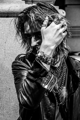 A person with an intense gaze is partially obscured by their hand, adorned with rings and bracelets. The individual wears a leather jacket and has long, textured hair, giving off a rock or punk aesthetic. Chains and jewelry add to the edgy look, and the photograph's black-and-white filter accentuates the mood.